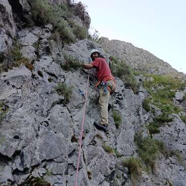 Un escalador, provisto de casco y arnés, asciende por una pared rocosa, utilizando cuerdas y equipo de seguridad. El entorno natural se caracteriza por un terreno accidentado y una vegetación frondosa.