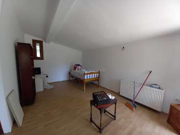This is a sparsely furnished room with a single bed, a wooden cabinet, a small table, a broom, radiator, and light wooden flooring.