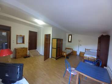 Esta imagem mostra um quarto espaçoso, mobiliado de forma simples, com piso de madeira, camas, uma escrivaninha, cadeiras, um guarda-roupa, prateleiras, uma cômoda e uma luminária.