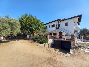 Uma casa branca de dois andares com telhado de telhas, escada externa e jardim. Cercada por árvores, plantas e um pátio aberto sob um céu azul claro.