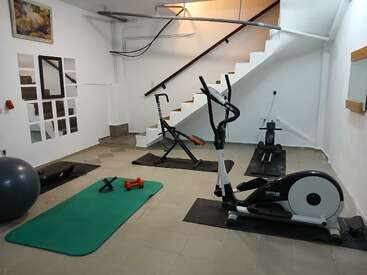 This image shows a small home gym with exercise machines, a green yoga mat, dumbbells, a stability ball, mirrors, and a staircase on a tiled floor.