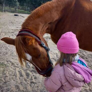 A young child in a pink hat and jacket gently interacts with a brown horse outdoors, showing a tender moment of connection and affection between them.