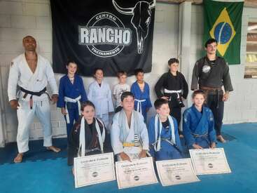 A group of children and two adults in martial arts uniforms pose for a photo. The children hold certificates. Behind them are banners and a Brazilian flag.
