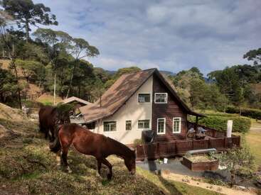 Two horses graze on a grassy hillside in front of a charming countryside house with a sloped roof, surrounded by trees, garden, and a wooden deck.