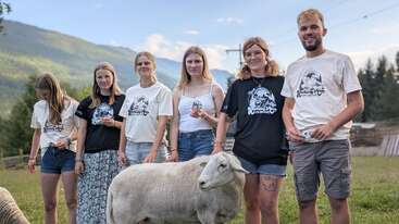 Six jeunes gens se tiennent à l'extérieur avec un mouton, souriant et tenant des objets. Ils portent des t-shirts assortis, entourés de collines vertes, d'arbres et d'un ciel bleu clair.