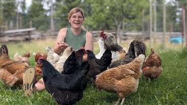 Une femme souriante est assise sur de l'herbe, entourée de plusieurs poules et d'un coq dans un décor de ferme, profitant du plein air et interagissant avec les oiseaux.