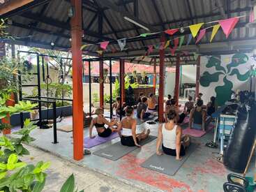 Um grupo de pessoas pratica ioga junto em esteiras num pavilhão ao ar livre decorado com bandeirinhas coloridas, cercado por plantas e luz natural, criando uma atmosfera tranquila.