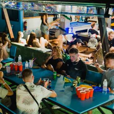 Um grupo animado de jovens se reúne em torno de uma mesa com bebidas, copos vermelhos e petiscos. Outros relaxam por perto; o clima é descontraído e festivo.