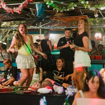 Uma animada festa em recinto fechado, com pessoas a rir, a cantar e a dançar sob decorações festivas. Duas mulheres estão em cima de uma plataforma, enquanto outras estão sentadas e apreciam o ambiente.