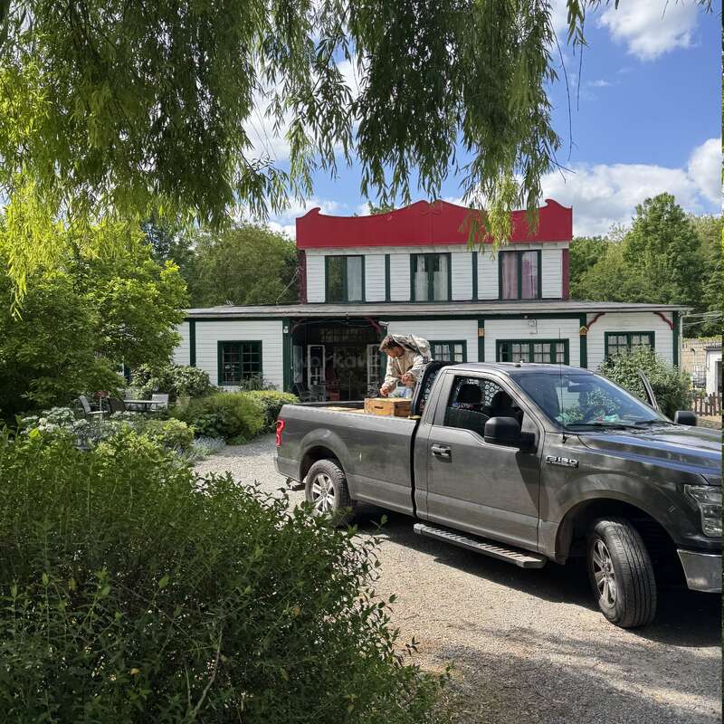 Eine Person entlädt Gegenstände aus einem grauen Pickup, der vor einem charmanten, zweistöckigen Haus inmitten von Grün unter einem sonnigen blauen Himmel geparkt ist.