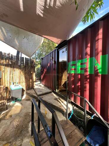This image shows a shaded outdoor area with bamboo fencing, a red shipping container with "GREEN" written on it, ramps, a trash bin, and trees overhead.