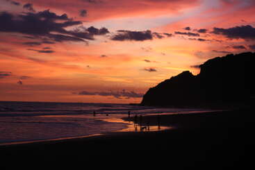 A stunning sunset paints the sky in shades of orange, pink, and purple above a beach, with silhouettes of people and cliffs along the shoreline.