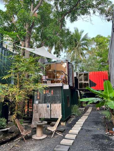 This image shows a rustic outdoor scene with shipping container buildings surrounded by lush greenery, trees, wooden chairs, a small round table, and a stone path.
