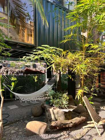 Modern container home with large windows, surrounded by lush tropical plants. A white hammock hangs in the shade, alongside a wooden chair and stone table.