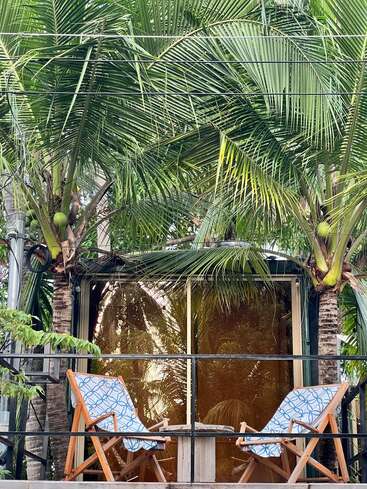Two patterned lounge chairs and a small table sit on a balcony, surrounded by lush green palm trees, creating a peaceful tropical relaxation spot.