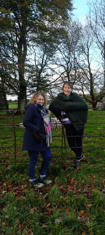 Zwei Frauen stehen lächelnd an einem Metallzaun auf einer Wiese, warm gekleidet in Mäntel und Schals, umgeben von Herbstbäumen und gefallenen Blättern.