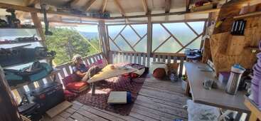 A cozy, rustic wooden cabin interior with open shelves, a rug, cushions, low table, and makeshift kitchen. A person relaxes, reading, surrounded by nature outside.