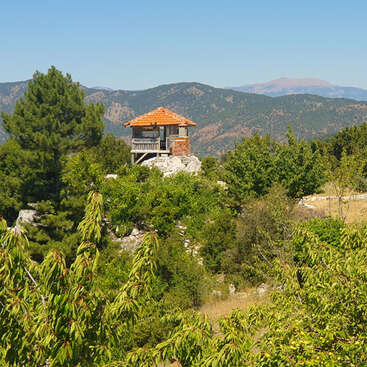 A small rustic house with a red-tiled roof stands atop a rocky hill, surrounded by lush green trees and overlooking distant forested mountains under a clear sky.