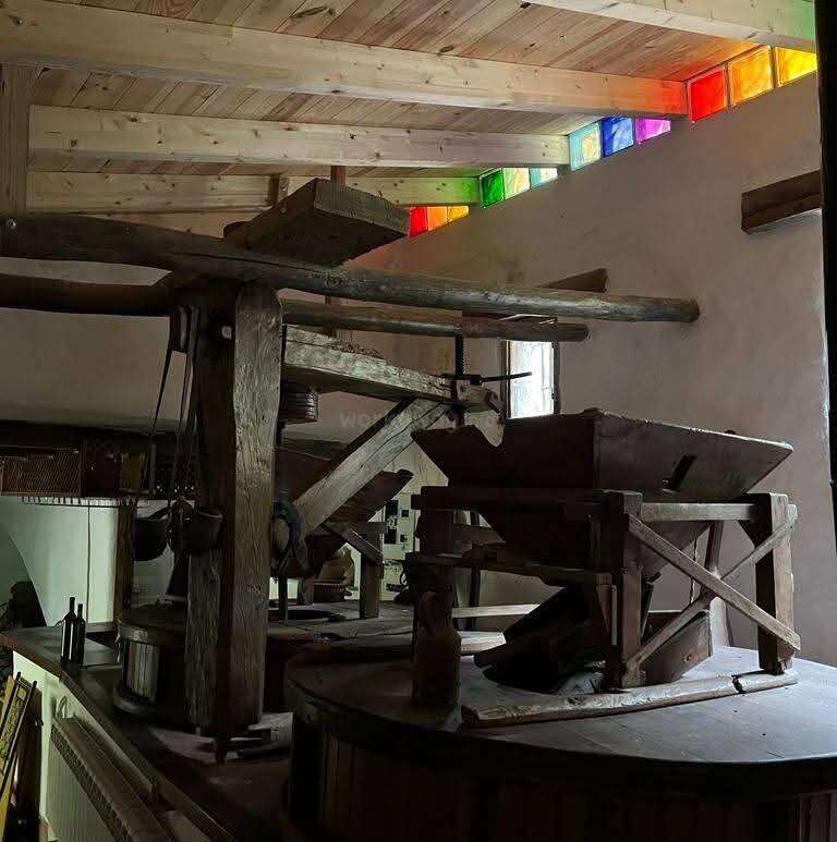 Esta imagem mostra uma antiga máquina de moagem de madeira em um cômodo rústico. Vitrais coloridos iluminam o espaço, criando uma mistura de tradição e beleza.
