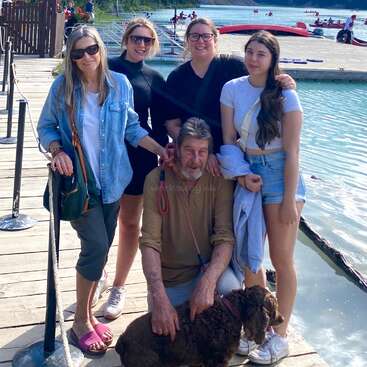 A imagem mostra uma família de cinco pessoas posando em um cais, com um cachorro a seus pés, em um lago sereno e um cenário montanhoso.
