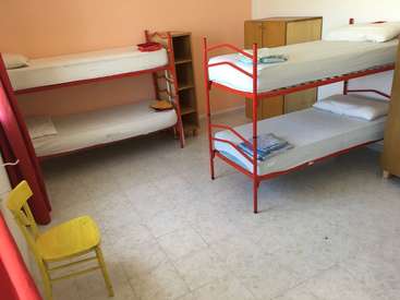 A imagem mostra um quarto com dois beliches, uma cadeira amarela e um armário, com roupas de cama brancas e molduras vermelhas, em uma parede cor de pêssego e piso de ladrilho.