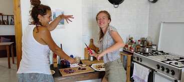 A imagem mostra duas mulheres cozinhando em uma cozinha, uma usando uma camisa branca e a outra usando um vestido branco, ambas de pé atrás de uma mesa com uma tigela de comida.