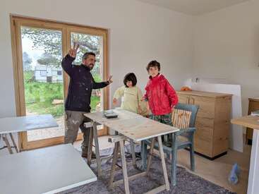 A man and two children are painting wooden boards inside a room near large windows. The man flashes a peace sign, and everyone seems engaged in work.
