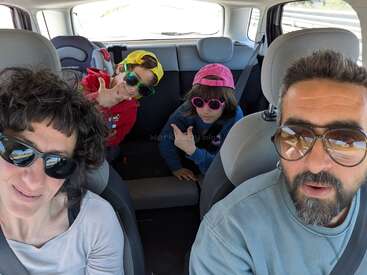 A family of four, all wearing sunglasses, poses for a selfie inside a car. The children in the back make playful hand gestures and wear colorful caps.