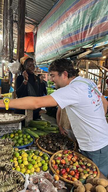 Un homme en chemise blanche sélectionne des produits sur un étal de marché animé. Autour de lui, des paniers de fruits et légumes divers, et un vendeur qui observe.