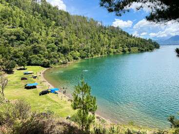 Une belle scène au bord d'un lac avec une eau bleue claire, des collines vertes et boisées, des gens qui se détendent sous des tentes, des voitures garées à proximité et un ciel ensoleillé.