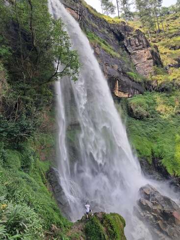 Una imponente cascada se precipita por un acantilado rocoso rodeado de una exuberante vegetación. Una persona se encuentra cerca de la base, capturando con su cámara las impresionantes vistas.