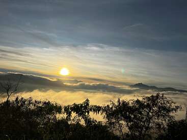 Das Bild zeigt eine ruhige Sonnenaufgangs- oder Sonnenuntergangsszene mit einer strahlenden Sonne, die durch die Wolken blickt, umgeben von einer nebligen Landschaft und silhouettierten Bäumen im Vordergrund.
