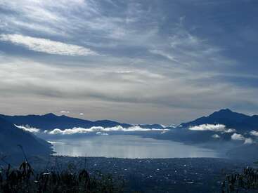 La imagen representa una serena escena lacustre con montañas al fondo, rodeada de nubes y un cielo azul, que evoca una sensación de belleza natural y tranquilidad.