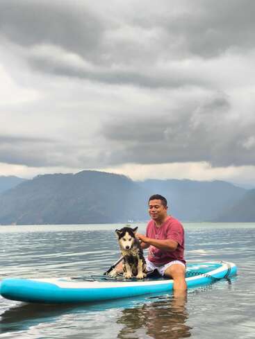 Um homem está sentado em uma prancha azul com um cão husky em um lago calmo, cercado por montanhas sob um céu nublado e cinzento. Uma aventura tranquila ao ar livre.
