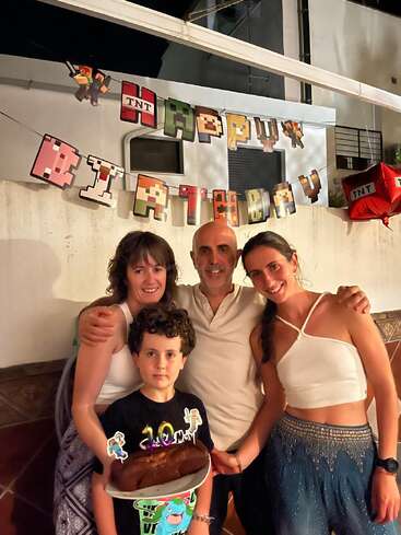 A family celebrates a child’s birthday with a Minecraft-themed banner in the background. The boy holds a chocolate cake, surrounded by smiling adults in warm lighting.