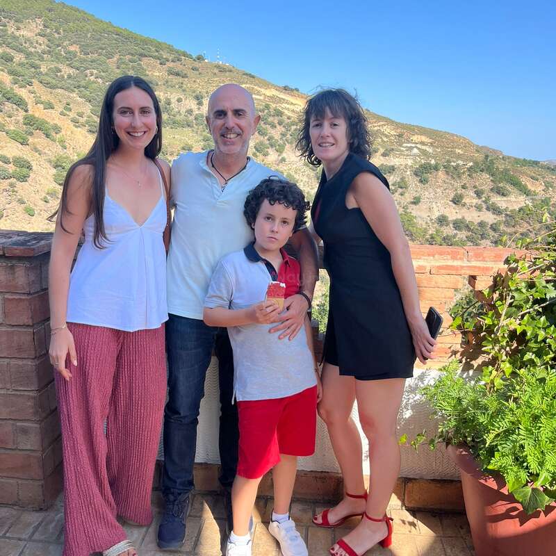 Four people pose together outdoors on a sunny day, with hills and greenery in the background. They look happy, enjoying the view and each other’s company.