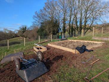 A rural garden scene shows raised beds being prepared for planting. Tools, an overturned wheelbarrow, and bare trees can be seen under a clear blue sky.