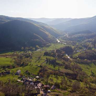 A imagem retrata uma paisagem rural serena com colinas onduladas, vegetação exuberante e uma pequena aldeia, em um céu claro e ensolarado.
