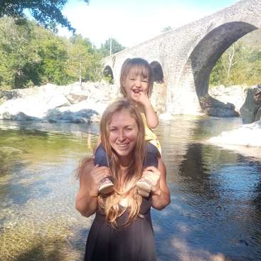 La imagen muestra a una mujer con un niño a hombros, de pie en aguas poco profundas, con un puente de piedra y una exuberante vegetación de fondo.