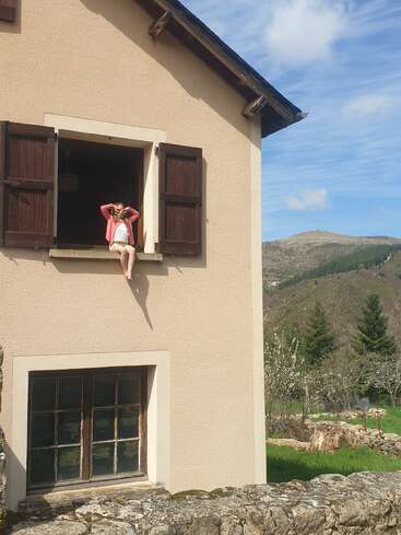 Uma garota está sentada em uma janela aberta de uma casa bege com persianas de madeira, relaxando, com montanhas, árvores verdes e um céu claro ao fundo.