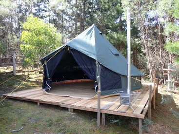 Ein großes grünes Zelt steht auf einer hölzernen Plattform in einem bewaldeten Gebiet. Das Zelt hat einen offenen Eingang und einen Metall-Holzofen draußen.