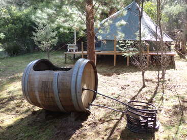 Im rustikalen Außenbereich steht ein großer Holzfass-Whirlpool mit Metallspulenheizung in der Nähe einer Zelthütte, die von Bäumen und natürlichem Grün umgeben ist.