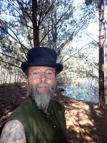Un hombre barbudo con gafas y sombrero de copa negro se encuentra en un bosque soleado cerca de un pequeño estanque, rodeado de árboles altos y naturaleza.