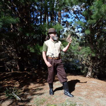 A bearded man in vintage explorer attire stands in a sunlit forest, observing a large pinecone. He wears a hat, boots, and brown trousers, surrounded by trees.