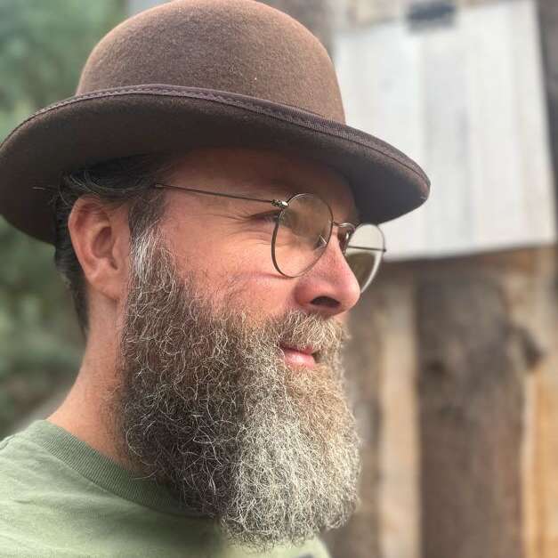 Ein bärtiger Mann mit einer runden Brille und einer braunen Melone blickt zur Seite. Er hat einen ruhigen Gesichtsausdruck und steht im Freien in der Nähe von Holz.