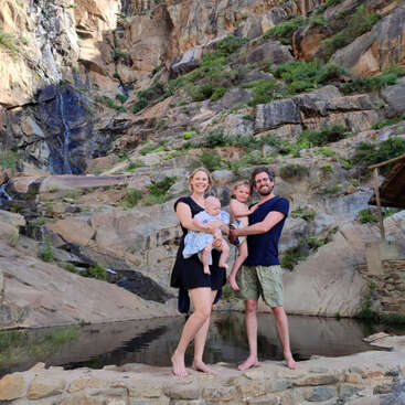 A imagem mostra uma família de quatro pessoas em pé sobre rochas perto de um pequeno lago, com um penhasco rochoso e uma cachoeira ao fundo, em um cenário natural ao ar livre.