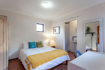 A imagem mostra um quarto aconchegante com uma cama, mesa e cadeiras, com um esquema de cores quentes e uma grande janela, evocando uma sensação de conforto e relaxamento.