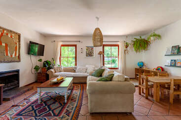 A imagem mostra uma sala de estar aconchegante com lareira, com uma mesa e cadeiras de madeira, uma estante e uma grande janela, exalando calor e conforto. A sala é bem iluminada e convidativa.