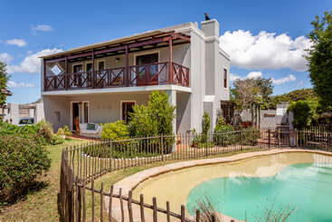 A imagem mostra uma casa com um grande quintal, com uma piscina e uma área de pátio, cercada por árvores e arbustos, com um céu claro ao fundo.