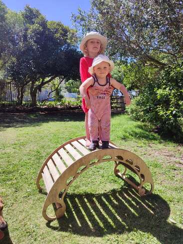 A imagem mostra duas crianças em pé em uma estrutura de madeira de um playground em uma área gramada, cercada por árvores e arbustos em um dia ensolarado.